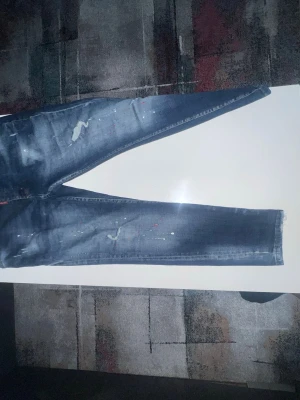  Dsq Mörkblå slitna  jeans - Säljer ett par mörkblå s jeans med slitningar och färgstänk för en cool, trendig look. Jeansen har smal passform och detaljerade slitningar på låren och knäna. Perfekta för dig som gillar streetstyle och vill sticka ut.