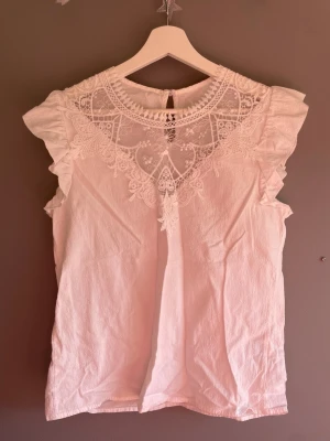 Vit blus med spetsdetaljer - Söt vit blus med volangärmar och detaljerad spets runt halsringningen. Blusen är ärmlös med romantisk känsla och har en liten knapp i nacken. Perfekt till jeans eller kjol för en fräsch look.