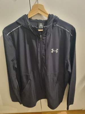 Svart jacka från Under Armour - Svart jacka från Under Armour med dragkedja framtill och vit logga på bröstet. Snygga detaljer med text längs axlarna och praktiska fickor. Tillverkad i ett lätt och sportigt material. Fast pris 525 kr.