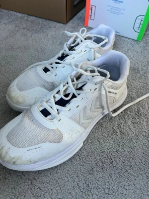 Vita Hummel sneakers med meshdetaljer - Snygga vita sneakers från Hummel med klassiska pilar på sidan och meshpaneler för extra ventilation. Skorna har vita snören, låg profil och detaljer i syntetmaterial. Perfekta för dig som gillar sportig och clean stil.