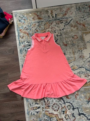 Rosa ärmlös klänning Holly & White - Söt rosa ärmlös klänning från Holly & White med pikékrage och knappar framtill. Klänningen har volang nertill och vita ränder på krage och ärmöppningar. Perfekt för varma dagar och enkel att styla.