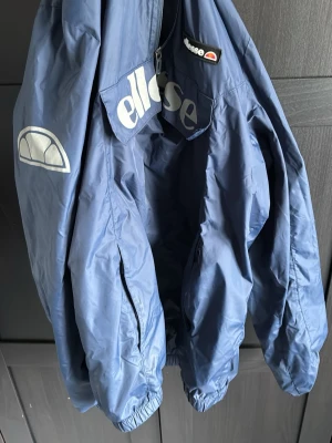 Blå vindjacka från Ellesse - Snygg blå vindjacka från Ellesse med stora logotryck på bröstet och ärmen. Jackan har dragkedja framtill, elastiska muddar och en avslappnad passform. Perfekt för att ge din outfit en sportig touch.