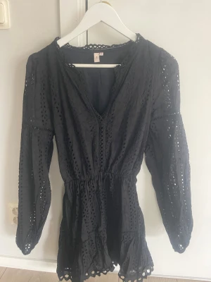 Sommarklänning från Nelly - Säljer en svart klänning från Nelly med v-ringning och långa ärmar. Klänningen har fina spetsdetaljer och broderade hålmönster, samt resår i midjan för en snygg passform