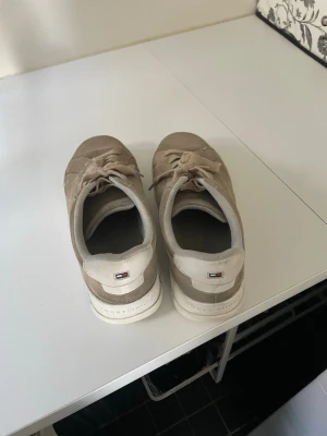 Beige sneakers från Tommy Hilfiger - Snygga beige sneakers från Tommy Hilfiger med ovandel i mocka och klassisk låg siluett. Skorna har vita sulor, diskret logga bak och matchande beige snörning. Perfekta för en clean och stilren look.