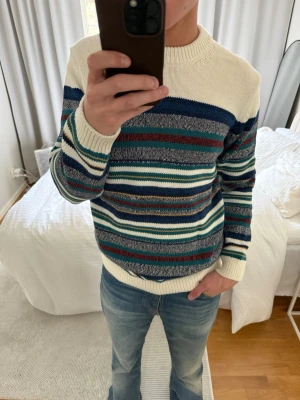 Randig stickad tröja från GANT - Säljer en stickad tröja från GANT i storlek M. Tröjan har krämvit bas med breda och smala ränder i blått, grönt, rött och grått. Rund halsringning och ribbstickade muddar. Perfekt för dig som gillar klassiska och färgglada plagg.