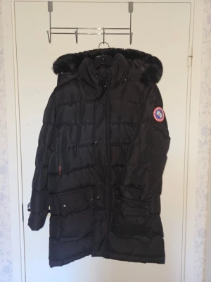 Svart dunjacka från Canada Goose - Snygg svart dunjacka från Canada Goose med huva och pälskant. Jackan har en längre modell, dragkedja framtill, två stora fickor och klassisk logotyp på ärmen. Perfekt för kalla vinterdagar och riktigt varm tack vare dunfyllningen.