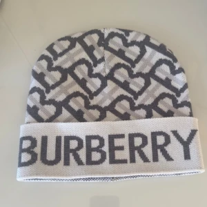 Gråvit Burberry mössa med logga - Säljer en snygg gråvit mössa från Burberry med stort logotyp-tryck framtill och ett mönster av sammankopplade B:n över hela mössan. Mössan har uppvikt kant och är stickad i mjukt material som känns lyxigt och trendigt.