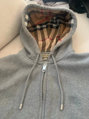 Grå hoodie från Burberry med dragkedja - Snygg grå hoodie från Burberry med klassiskt rutigt foder i huvan. Dragkedja framtill, justerbara snören och diskret broderad logga på bröstet. Perfekt för en stilren och avslappnad look.