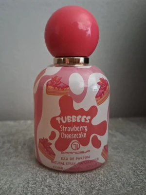 Tubbees Strawberry Cheesecake parfym - Söt parfymflaska från Grandeur med doften Tubbees Strawberry Cheesecake. Flaskan är dekorerad med rosa och vita färger samt bilder på jordgubbscheesecake. Rund rosa kork och guldiga detaljer ger en lekfull och unik look. Perfekt för dig som gillar söta dofter.