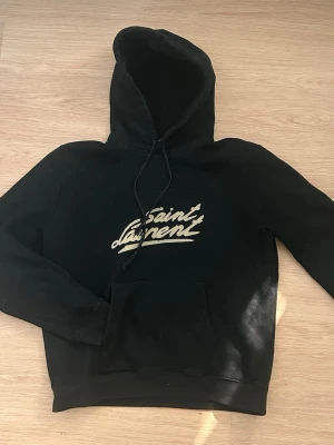 Svart Saint Laurent hoodie - Svart hoodie från Saint Laurent med vit logga på bröstet i snyggt skriven stil. Klassisk känguruficka framtill och justerbar dragsko i huvan. Perfekt för dig som gillar streetwear och exklusiva märken.