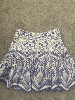 Vit och blå mönstrad kjol från Zara - Kort kjol från Zara i vitt och blått med dekorativt mönster och volangkant. Kjolen har knappar framtill och är tillverkad i ett lätt bomullstyg, perfekt för varmare dagar. Snygg och trendig design som sticker ut.