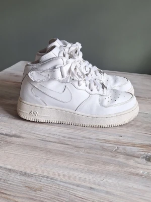 Nike Air Force 1 vita midsneakers stl 38,5 - Klassiska vita Nike Air Force 1 midsneakers i läder med snörning och kardborreband vid ankeln. Swoosh-logga på sidan och Nike Air-brodyr på hälen. Tjock sula med AIR-dämpning och perforeringar på tån för ventilation.
