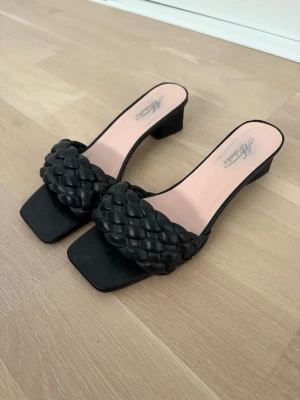 Svarta flätade slip-in sandaler - Säljer ett par svarta slip-in sandaler köpta i Frankrike med bred, flätad rem och fyrkantig tå. Sandalerna har en låg, stabil klack och innersula i ljusrosa. Perfekta för dig som gillar trendiga och bekväma skor med en modern twist.