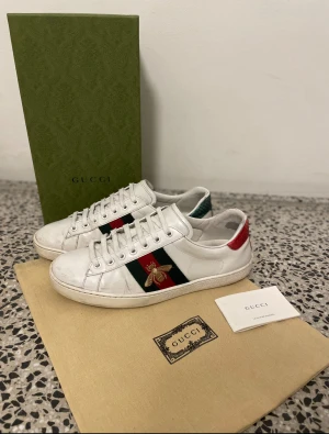 Gucci Ace sneakers med broderad bi - Gucci Ace sneakers i vitt skinn med klassiska gröna och röda ränder på sidan och broderad guldfärgad bi. Baksidan har metallicröd och metallicgrön detalj med textur. Platt sula, rund tå och snörning. Kommer med originalkartong och dustbag.