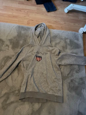 Grå hoodie från GANT med emblem - Snygg grå hoodie från GANT med broderad logga och ett emblem med stjärnor och ränder på bröstet. Klassisk huva och känguruficka framtill. Perfekt för en chill och sportig stil. Mjuk bomullsblandning som är skön att ha på sig.