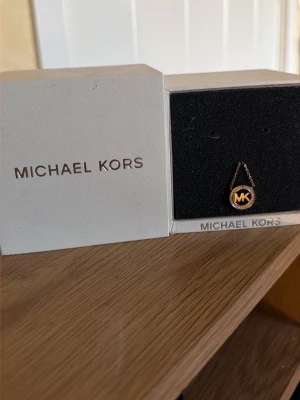 Guldigt halsband från Michael Kors - Snyggt halsband från Michael Kors i guldton med rund hängberlock och MK-logga. Kedjan är tunn och elegant, och berlocken har glittrande detaljer runt loggan. Perfekt accessoar för att lyfta din outfit.