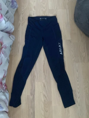 Marinblå ridtights från Ariat - Snygga marinblå ridtights från Ariat med hög midja och vitt logotryck på benet. Tightsen har ventilerande meshpaneler längs benen för extra komfort och rörelsefrihet. Perfekta för träning eller ridning. Har lite hål då de är väll använda. Storlek 152 men jag som har mer åt 36 hållet kommer i dem bara att de har blivit för korta för mig. 