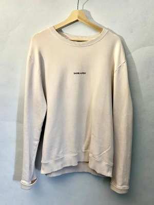 Saint Laurent Archive Logo Sweatshirt - Vit/krämfärgad tröja från YSL. Denna modell har en distressed look från fabrik, vilket betyder att både muddar och print har ”slitningar”. Sällsynt modell i riktigt fint skick. Det ända lilla flawet är att tröjan saknar neck tag, annars är den som ny.