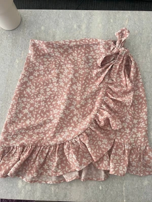 Rosa kjol med volanger - Superfin rosa kjol med vita blommor och volangkant. Kjolen har en knytning i sidan och fina ryschdetaljer längs kanten. Perfekt för sommaren och och semestrar. Materialet känns lätt och luftigt med bra kvalitet.