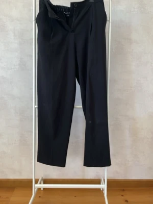 Svarta kostymbyxor med resår - Säljer ett par svarta kostymbyxor med rak passform och resår i midjan. Byxorna har pressveck fram och två bakfickor. Perfekta för en clean och stilren look. Materialet känns mjukt och följsamt.
