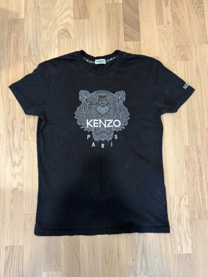  Kenzo t-shirt  - Svart t-shirt från Kenzo med ikoniskt tigertryck och texten 'KENZO PARIS' på bröstet. Klassisk rund halsringning och korta ärmar. Märkeslogga även på ärmen. Perfekt för dig som gillar streetwear och vill sticka ut.
