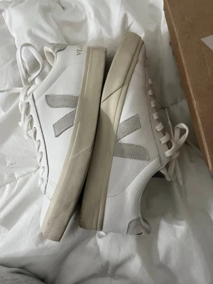 Vita Veja sneakers med grå detaljer - Säljer dessa snygga vita sneakers från Veja med grå V-logga på sidorna. Passar perfekt nu till sommaren. De är i storlek 40 och är endast använda en gång! Säljer pga de tyvärr är för stora. Skriv om du har några funderingar💗 