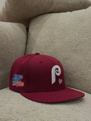 Vinröd Phillies keps från New Era - Säljer en vinröd snapback-keps från New Era med Phillies-logga i vitt framtill och broderad detalj med hjärta. På sidan finns ett broderat märke för World Series 1980. Klassisk rak skärm och broderade ventilationshål. Perfekt för dig som gillar sportig streetstyle.