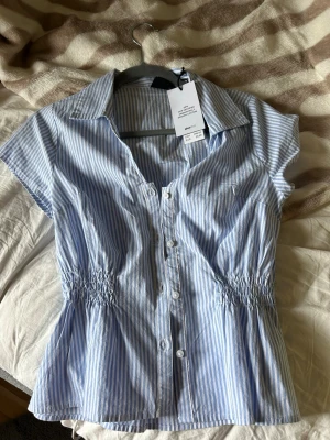 Blårandig smockblus från Gina Tricot - Snygg blus med blå och vita ränder, smockad midja och korta ärmar. Klassisk krage och knäppning framtill. Tillverkad i mjuk bomull med en luftig och avslappnad passform. Perfekt till jeans eller kjol för en fräsch look.