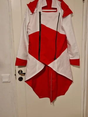 Assasins creed Cosplay jacka small - Unik cosplayjacka i rött och vitt med asymmetrisk design, stor huva och två svarta dragkedjor framtill. Jackan har långa ärmar, röda detaljer på ärmslut och en längre bakdel. Perfekt för maskerad eller cosplayevent!