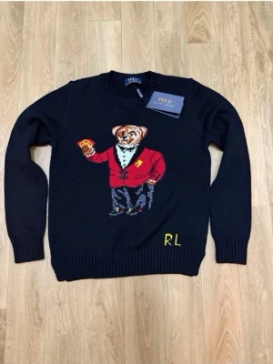 Mörkblå stickad tröja Polo Bear - Mörkblå stickad tröja från Polo Ralph Lauren med ikoniska Polo Bear-motivet framtill. Björnen har röd kavaj och håller spelkort. RL-broderi i gult nedtill. Tröjan har rund hals och ribbade muddar. Perfekt för dig som gillar statement-plagg.