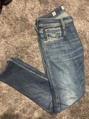Blå slim fit jeans från Replay - Säljer ett par blå slim fit jeans från Replay med snygga slitningar och klassisk femficksdesign. Jeansen har en ljus tvätt med markerade sömmar och är tillverkade i mjuk denim som sitter skönt. Perfekta för dig som gillar en modern och avslappnad stil.
