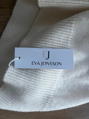 Vit polotröja från Eva Jonsson - Säljer en stilren vit-beige stickad polotröja från Eva Jonsson. Tröjan har ribbade muddar vid ärmslut och nederkant samt en hög polokrage. Perfekt för dig som gillar klassisk och enkel stil.