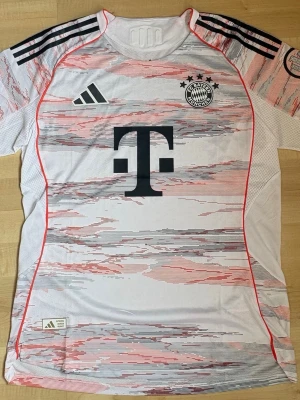FC Bayern München 25/26 away - Snygg FC Bayern München matchtröja från Adidas i vitt med röda och gråa grafiska detaljer. Tröjan har klubbmärke och Adidas-logga på bröstet, samt Telekoms sponsortryck. Svarta ränder på axlarna och texten 'FC Bayern München' på ryggen. Tillverkad i lätt och ventilerande material.
