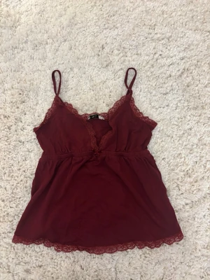 Vinröd spetslinnetopp med smala band - Snygg vinröd linnetopp med tunna axelband och spetsdetaljer längs urringning och nederkant. Toppen har en v-ringad form och är tillverkad i mjukt material som känns skönt mot huden. Perfekt för att addera en romantisk touch till din outfit.