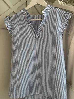 Blårandig blus med volangärm - Snygg blus med tunna blå och vita ränder, V-ringad hals och volangdetaljer vid ärmarna. Blusen är i ett lätt och luftigt material, perfekt för sommaren. Passar till både jeans och kjol för en fräsch look.