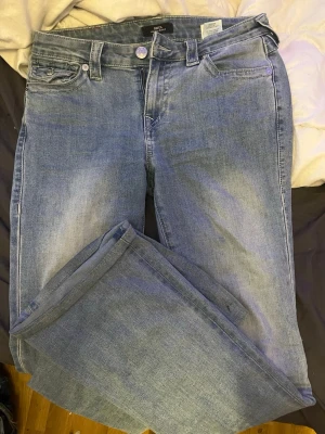 Blå jeans från True Religion - Säljer ett par klassiska blå jeans från True Religion med lite utsvängda ben och snygga detaljerade bakfickor med knappar. Jeansen har fem fickor, bälteshällor och en ljus tvättad look. Perfekta för dig som gillar streetwear och vill ha ett par jeans med ikonisk logga. Använda 1 gång och säljer då dom är i fel storlek för mig.