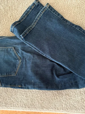 Mörkblå raka jeans - Säljer ett par mörkblå jeans med rak passform och klassisk femficksdesign. Jeansen har kontrastsömmar och är tillverkade i denim, vilket ger en tidlös look. Perfekta för dig som gillar enkel och stilren design.