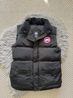 Svart dunväst från Canada Goose (M) - Snygg svart dunväst från Canada Goose med klassisk röd och vit logga på bröstet. Västen har hög krage, två stora fickor framtill och stängs med både dragkedja och tryckknappar. Perfekt för lager-på-lager och riktigt varm tack vare dunfyllningen.