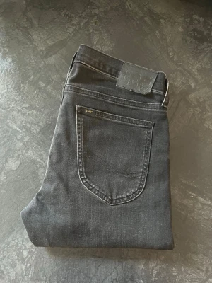 Lee Slim Fit Jeans - Snygga svarta slim fit jeans från Lee med klassisk femficksdesign och diskret Lee-logga på bakfickan. Jeansen har en smal passform och är tillverkade i ett mjukt denim-material som ger en bekväm känsla. Perfekt för dig som gillar en stilren look. Storlek W29 L30. 