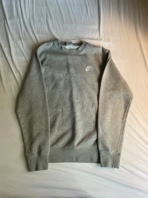 Grå sweatshirt från Nike - En klassisk grå sweatshirt från Nike med broderad logga på bröstet. Tröjan har rund halsringning, långa ärmar och ribbade muddar. Perfekt för en avslappnad och sportig stil.