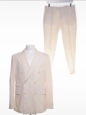 Vit dubbelknäppt kostym från Oscar Jacobsson  - En elegant kostym från Oscar Jacobsson i en stilren cremefärg/benvit. Tillverkad i 100% ull vilket ger en exklusiv känsla och fin passform. Perfekt för fest, bröllop eller andra uppklädda tillfällen.  Kostymen består av en dubbelknäppt kavaj och byxa, båda i mycket gott skick. Båda delar är tvättade av kemtvätt och har inte använts efter de. Klassisk design som aldrig går ur tiden.  Storlek: 50 på både kavaj och byxor  Skick: Mycket bra  Hör gärna av dig vid frågor! 😊