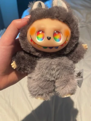 Grå plysch nyckelring från The Monsters - Söt och fluffig Labubu nyckelring från The Monsters, formad som en liten figur med grå päls och silvriga detaljer på öronen. Nyckelringen har en svart ring och ett band med logotypen. Perfekt att hänga på väskan eller nycklarna. ( labubun är fejk ) 