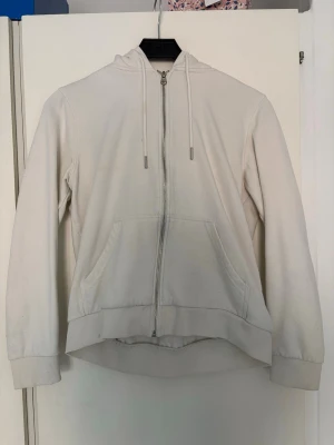 Vit hoodie med dragkedja från H&M - Snygg vit hoodie från H&M Basics med dragkedja framtill och två stora fickor. Klassisk modell med huva och dragsnören. Perfekt för en chill och avslappnad stil. Tillverkad i mjukt bomullsmaterial som är skönt mot huden.