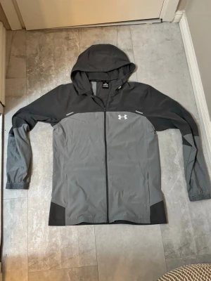 Grå vindjacka från Under Armour - Snygg grå och svart vindjacka från Under Armour med huva och hel dragkedja. Jackan har logga på bröstet och stor text på ryggen. Tillverkad i lätt, vindtätt material och har elastiska muddar. Perfekt för blåsiga dagar och sportiga outfits.   Den är använd endast ett par gånger så den är i typ nyskick          RESERVERAD 