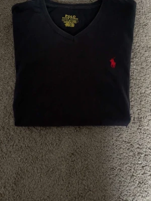 Svart v-ringad t-shirt Polo Ralph Lauren - Svart v-ringad t-shirt från Polo Ralph Lauren med röd broderad logga på bröstet. Slim fit modell i mjuk bomull som sitter snyggt och stilrent. Perfekt för dig som gillar klassisk och enkel stil med en touch av lyx.