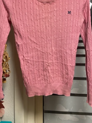 Rosa kabelstickad tröja från Morris - Snygg rosa kabelstickad tröja från Morris med rund halsringning och diskret broderad logga på bröstet. Tröjan har långa ärmar och ribbade muddar vid ärmslut och nederkant. Perfekt för dig som gillar klassisk preppy stil.