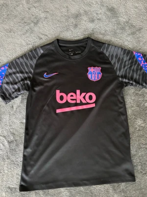 Svart FC Barcelona träningströja  - Snygg svart träningströja med FC Barcelona-logga och rosa detaljer. Tröjan har korta ärmar med zebramönster och blå/rosa inslag, samt rund halsringning. Tillverkad i lätt och ventilerande material, perfekt för fotbollsträning.