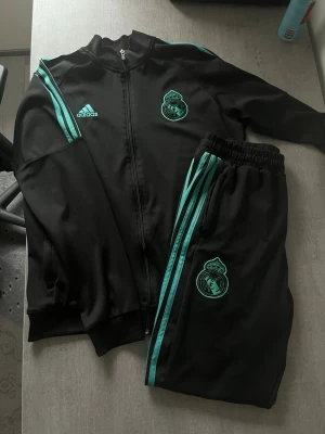 Real Madrid svart träningsdress Adidas - Säljer en svart träningsdress från Adidas med Real Madrid-logga på både jacka och byxor. Setet har turkosa detaljer och klassiska Adidas-ränder längs ärmar och ben. Jackan har dragkedja och fickor, byxorna har resår i midjan. Perfekt för fotbollsträning.