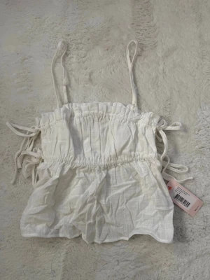 Vit linnetopp med knytband - Söt vit linnetopp med tunna axelband och knytband i sidorna. Toppen har en rynkad, elastisk midja och är tillverkad i ett lätt, luftigt material som passar perfekt till varma dagar. Enkel och stilren design som är lätt att matcha. helt ny köpt förra året men aldrig använd 