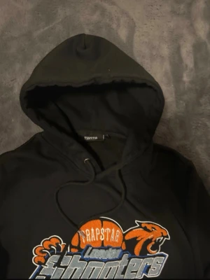 Trapstar London Shooters hoodie svart - Svart hoodie från Trapstar med stor broderad logga på bröstet där det står 'London Shooters' och en orange basketboll med pantermotiv. Klassisk huva med dragsko och mjukt material. Perfekt för dig som gillar streetwear och sportiga vibes.